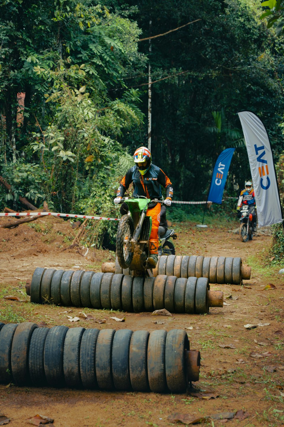 Dirt bike rider jumps over stacked tires on a muddy track.