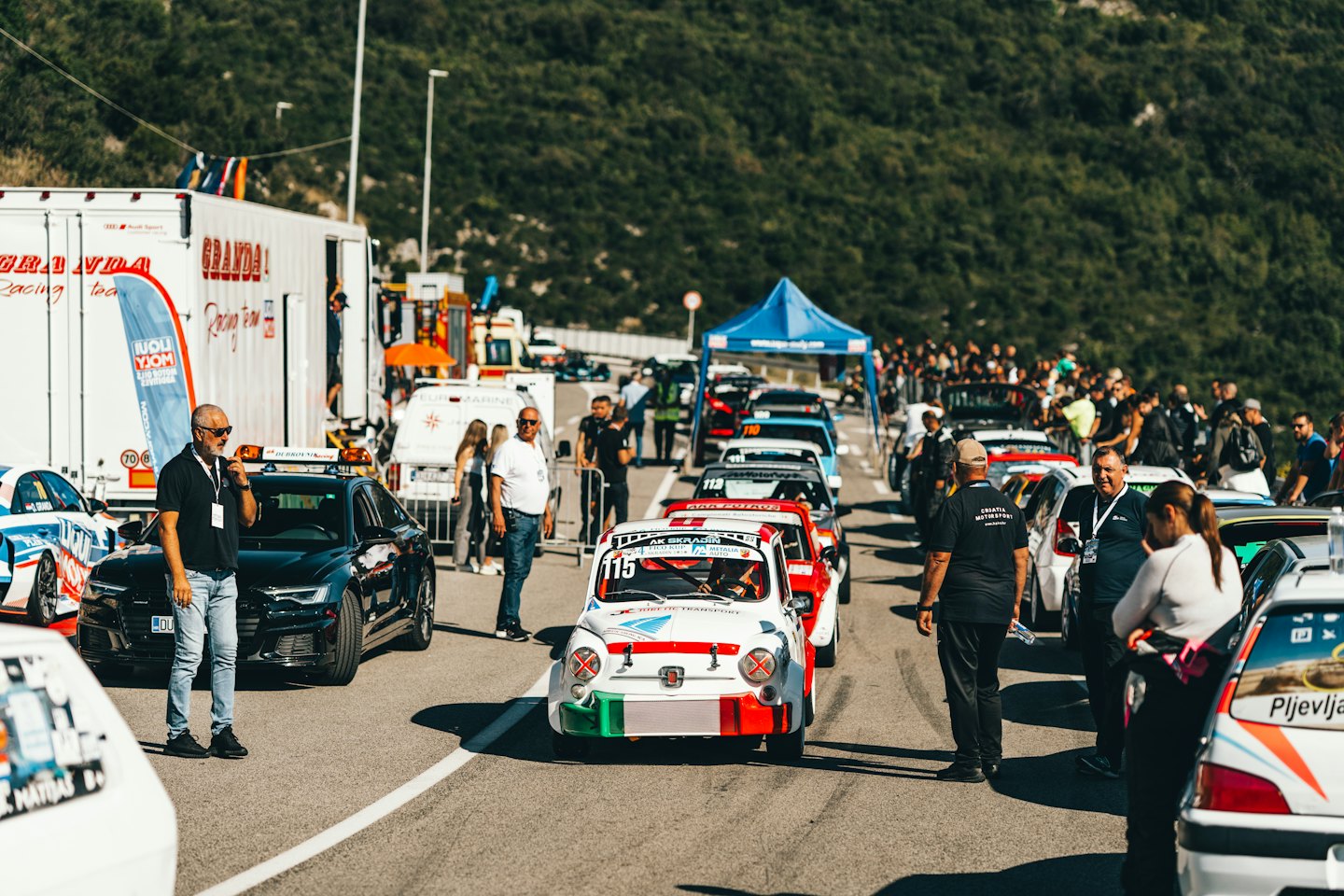 People and race cars gather on a road near mountains.